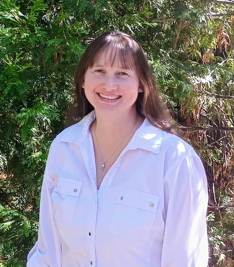 Cher Wolin, smiling, in a white shirt in front of green shrubs